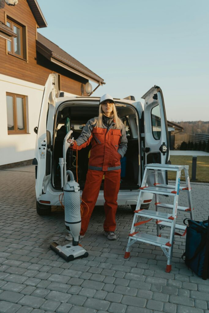 pexels photo 6196239 6196239 1 A cleaner in uniform stands beside a van with cleaning tools outside a modern house at sunset.